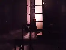 A blond woman with a turquoise bob wig, singing to a microphone, in front of two large door panels. Steam billows around her.