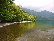 Photo couleur d'un lac à l'eau transparente. Au second plan, s'étend le littoral boisé du lac, sous un ciel chargé de nuages.