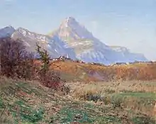 Sur un coteau aux couleurs automnales, un village avec un clocher est dominé en arrière-plan par une montagne.