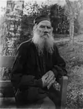 Photo noir et blanc d'un vieillard à grande barbe blanche, assis mains croisées sur un banc dans un jardin.