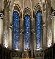 Photographie en couleurs d’une chapelle vue de face avec trois vitraux et de part et d'autre une mosaïque murale et en bas la statue de saint Jean.
