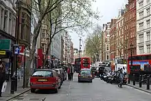 Une rue pavée bordée d'immeubles anciens rouges et blancs. Parmi la circulation routière se trouvent deux bus rouges à étage.