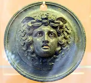 Dans un cercle, tête de Méduse entourée de cheveux, avec des serpents à la base du cou et des ailes en arrière de la tête. Bouche grande ouverte, yeux globuleux.