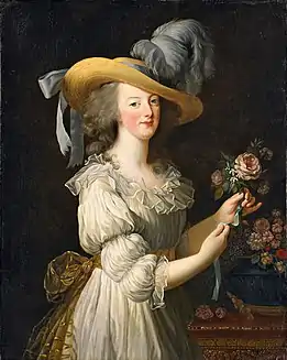 Portrait de trois-quarts d'une femme portant un grand chapeau de paille avec des plumes d'autruche bleu-gris, et une robe couleur crème avec un noeud doré à la ceinture dans le dos. Elle arrange un bouquet de fleurs roses.