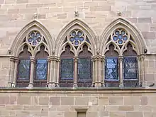 Photo couleur d'un groupe de trois fenêtres géminées en ogive double à colonnettes. Les baies sont munies de panneaux de bois dotés de vitraux en losange.