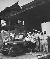 Photographie en noir et blanc d’une dizaine d’individus habillés en blanc autour d’une Jeep, devant un château.