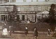 Photographie ancienne sur laquelle plusieurs enfants joue dans le jardin à l'arrière d'un manoir avec baie vitrée et véranda imposante.