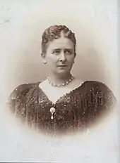 Photographie en buste de Marie les cheveux coiffés vers l'arrière, portant un rang de pierres précieuses entourant son cou et vêtue d'une robe décolletée sombre ornée d'un pendentif central