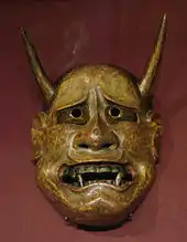 Masque non humain, jaunâtre, avec des cornes, de grandes canines, au regard implorant.