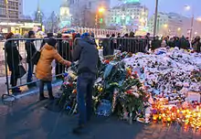 Plusieurs bougies et gerbes de fleurs sous la neige déposées au sol entourées de personnes.