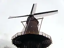 Photographie en couleurs de la partie supérieure d'un moulin à vent vue du dessous pointant vers le firmament d'un ciel nuageux.