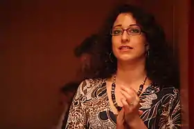 Portrait en buste d'une femme aux cheveux noirs et frisés, portant des lunettes de vue et un vêtement aux motifs végétaux blancs, noirs et marrons. Devant un fond rougeâtre, elle semble applaudir et regarder au loin.