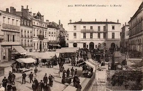 Photo ancienne de la place, un jour de marché. La place est bordée côté est par le théâtre, côté nord par la maison du Lyon (actuelle Société Générale), et côté ouest par l'ancien hôtel de ville