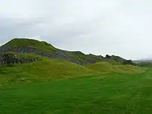Photo d'un paysage vallonné recouvert d'herbe sous un ciel nuageux. On distingue les ruines de murs de pierre grise