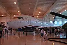 Photo prise à l'intérieur d'un hangar montrant l'avion endommagé exposé dans un musée. Des visiteurs sont présents tout autour et admire l'avion.