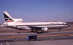 Photo couleur de l'avion, caractérisé par ses trois moteurs, dont deux sont situés sous ses ailes et le troisième sur sa queue. L'avion est peint en blanc et une fine ligne bleue sur le milieu et l'inscription Delta sur l'avant du fuselage. Il se trouve à une porte d'embarquement et des bâtiments de l'aéroport sont visibles à l'arrière-plan.