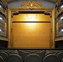 Un grand rideau doré ferme la scène. On distingue les fauteuils du parterre et les balcons.