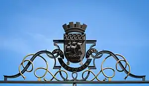 Écu sur fond de ciel bleu, sur un support de fer forgé aux formes courbes.