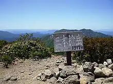 Photo couleur montrant une pancarte, plantée dans un sol gris pierreux partiellement recouvert de buissons verts, en bois annonçant l'altitude d'un sommet de montagne, une chaîne de montagne boisée au second plan et un ciel bleu en arrière-plan.