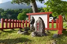 Photo couleur d'une stèle en pierre gravée d'une inscription en rouge et entourée d'autres pierres sur une étendue de verdure avec des arbres en arrière-plan.