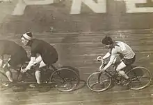Photographie en noir et blanc montrant un coureur cycliste derrière un tandem sur la piste d'un vélodrome.