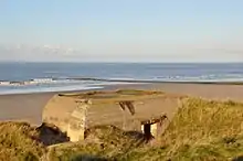 Photographie d'un bunker en ciment, à l'état de ruine, situé à l'abri d'une dune, au bord d'une plage.