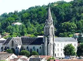 Photo représentant sur fond de verdure l'église Saint-Georges. Au centre, son clocher flèche et sur la gauche sa nef de laquelle ressort son transept proéminent. À droite du clocher, un long et haut bâtiment de trois étages situé derrière l'église donne l'impression d'une autre nef, presque symétrique.