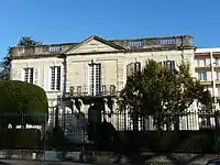 Bâtiment haut d'un étage et large de 5 hautes fenêtres, avec une rambarde sur son toit plat, précédée d'une terrasse et de sa grille de clôture.
