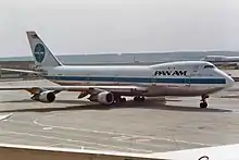 Photo d'un avion de Pan Am vu de trois quart face sur le tarmac gris d'un aéroport. L'avion, en gros plan, est stationné et entouré de bâtiments de faible hauteur et d'un autre avion et d'une forêt en arrière-plan.