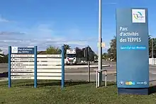 À gauche, un panneau indique le nom des entreprises du Parc d'activités des Teppes ainsi que leur direction. À droite se situe un totem annonçant la présence de la zone d'activités.