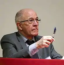 Photographie d'un homme assis vêtu d'un costume, portant des lunettes et tenant un stylo bille bleu dans sa main droite.