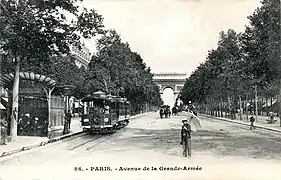 Photo noir et blanc d'une grande artère bordée d'arbres avec piétons, tramway, voitures à cheval, édicule à marquise ronde et au fond, l'arc de triomphe