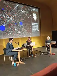 Projection d'une galaxie de liens en arrière plan, 3 femmes réparties sur une scène lisant du texte à tours de rôle sur leur ordinateur portable, dans un micro