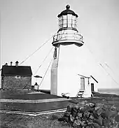 photo noir et blanc avec le phare, avec ses étais, au centre de l'image, sur la gauche une maison, au premier plan un muret en pierre sur la droite et la toiture de la citerne d'eau à gauche. En arrière-plan c'est majoritairement le ciel avec une petite vue sur la mer à droite