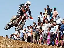 Une moto est en l'air, survolant de la terre. derrière, une foule de spectateurs.