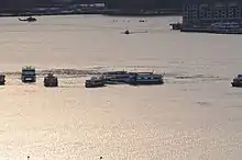 Photo prise au moment du coucher du soleil. L'avion est au centre de l'image, dans le fleuve tendant vers le jaune-orange dû à la réflexion du soleil couchant. Sur l'image, au moins sept ferries entourent l'avion ainsi qu'un hélicoptère en haut à gauche de l'image.