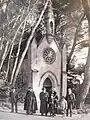 Plusieurs personnes dont un prêtre catholique en habit liturgique devant la chapelle du Plantier, entourée de grands pins.