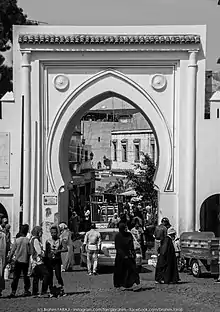 Photographie en noir et blanc d'une grande porte extérieure blanche aux formes orientales. Plusieurs personnes et véhicules entrent et sortent par la porte.