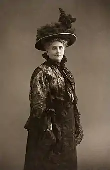 Gina krog debout, 3/4 profil regardant face caméra, robe, chapeau, légèrement souriante, fière.