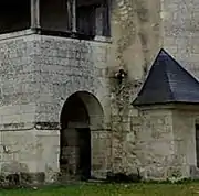 Photographie en couleurs d'une galerie en hauteur surmontant un angle de bâtiment ajourné d'une entrée cintrée, un proche à toiture triangulaire couvert d'un toit fait d'ardoises visible sur la droite.