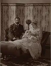 Devant un paravent aux motifs géométriques, Philippe en redingote, barbe sombre et portant des lunettes est assis auprès de Louise laquelle se tient sur une chaise longue, coiffée de dentelles, portant une robe claire et la tête inclinée vers son fils-nouveau-né dans ses bras.