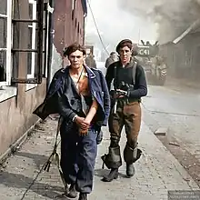 Photo colorisée montrant au premier plan un homme en uniforme, blessé, la veste ouverte, accompagné d'un homme portant un fusil d'assaut. Derrière eux, dans la rue d'un village, on voit de la fumée et un char d'assaut.