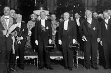 Photo en noir et blanc sur laquelle figurent notamment quatre individus en queue-de-pie se tenant debout