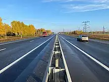 Photographie prise depuis le milieu de la route, avec deux voies de chaque côté, séparées par un séparateur. Du côté gauche, la route est bordée par des mélèzes de Sibérie orangés, car ils vont perdre leurs aiguilles, tandis qu'à droite des pylônes électriques s'élèvent sur un terrain déboisé.