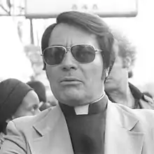 Photo en noir et blanc de Jim Jones, portant un vêtement de pasteur et des lunettes noires, devant une foule et un panneau qui indique Hotel.