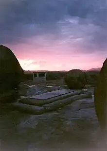Photographie d'un lever de soleil sur un paysage montagneux avec des arbres sur l'horizon. Au premier plan se trouve une pierre tombale rectangulaire tandis qu'un monument massif est visible quelques dizaines de mètres derrière.