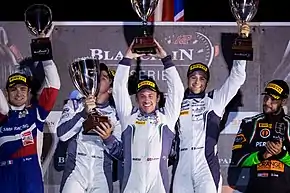 Photographie de cinq hommes sur un podium, vus de face, levant leurs trophées, dans la nuit.