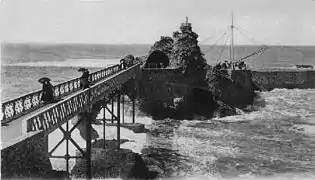 Photographie en noir et blanc d'un rocher sur la mer relié à la côte par une passerelle.