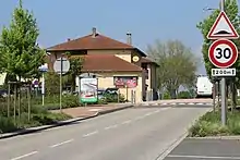Une route avec deux panneaux à droite dont un indique que la voie est limitée à 30 km/h. L'autre panneau indique la présence d'un ralentisseur. À gauche de la route se trouve un abri-bus devant un bâtiment jaune faisant office de restaurant.