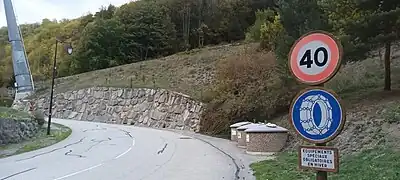 Photographie d'un panneau routier qui indique que la limitation kilométrique est de 40 km/h ; un équipement de pneus est nécessaire en hiver ; on voit les conteneurs à ordures pour le tri sélectif derrière, le tout depuis une route.